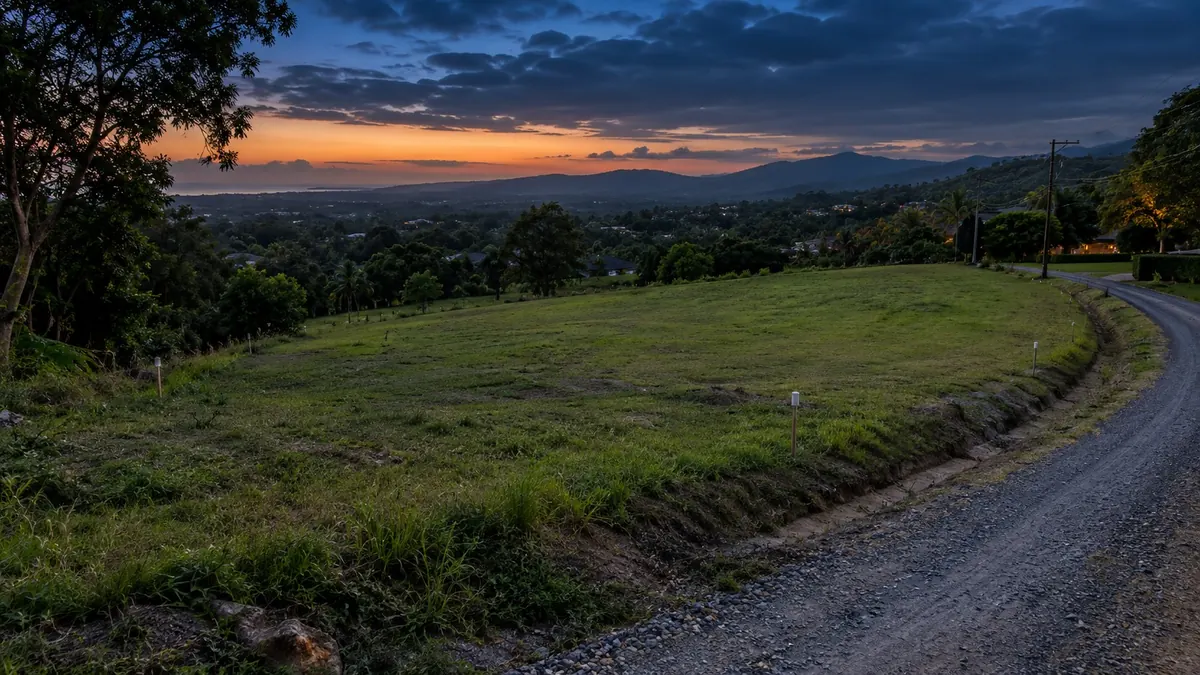 A hillside lot marked out with survey pegs beside a gravel road, the sea beyond at sunset — land, boundaries and title